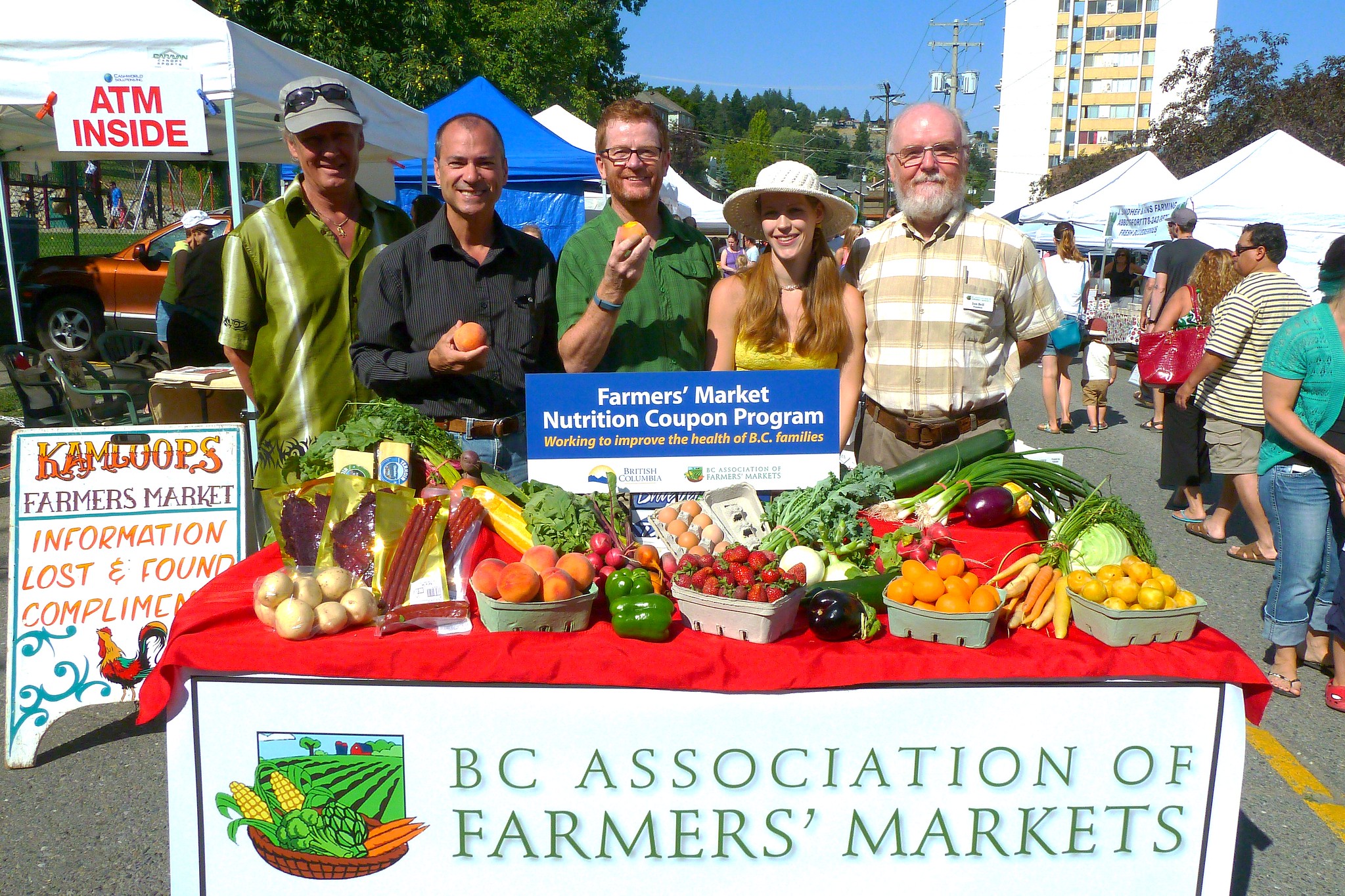 People smile around fresh produce and a sign saying &quot;BC Association of Farmers&#039; Markets.&quot;