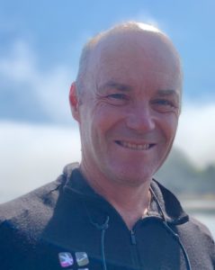 A balding man in a fleece jacket smiles in bright sunlight.