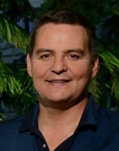 A man in a polo shirt smiles in front of a coniferous tree.