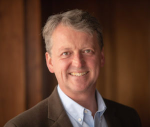 A middle-aged man in a blazer and collared shirt poses and smiles.