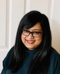 A woman in a turtleneck and glasses smiles in front of a white door.