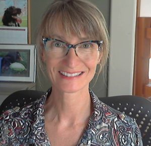 A blonde woman in a blouse and glasses smiles in an office chair.