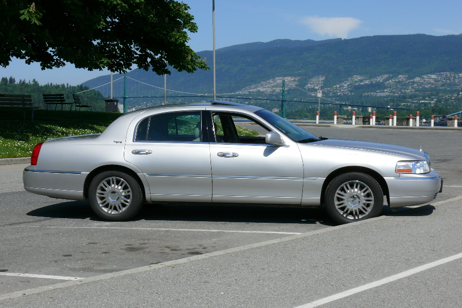 A silver car in a parking lot. A suspension bridge is in the background.