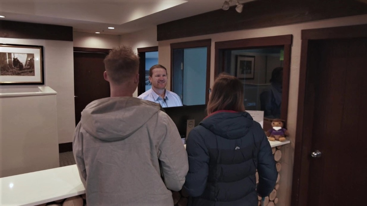 Two guests in winter coats talk to a hotel front desk employee.