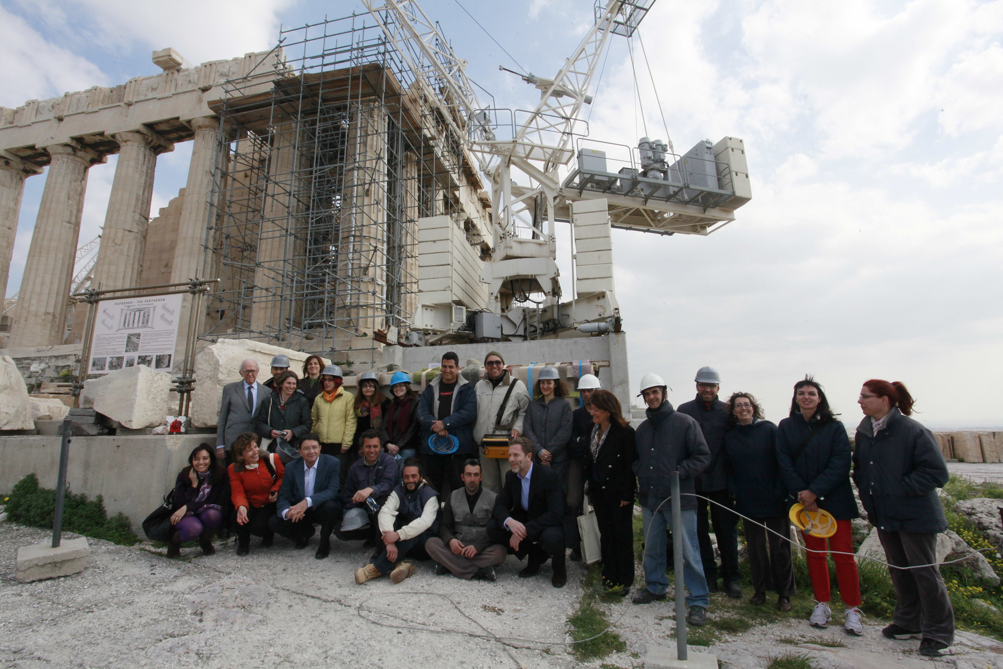 A group poses in front of ancient Greek ruins covered in scaffolding. Some people wear hard hats.