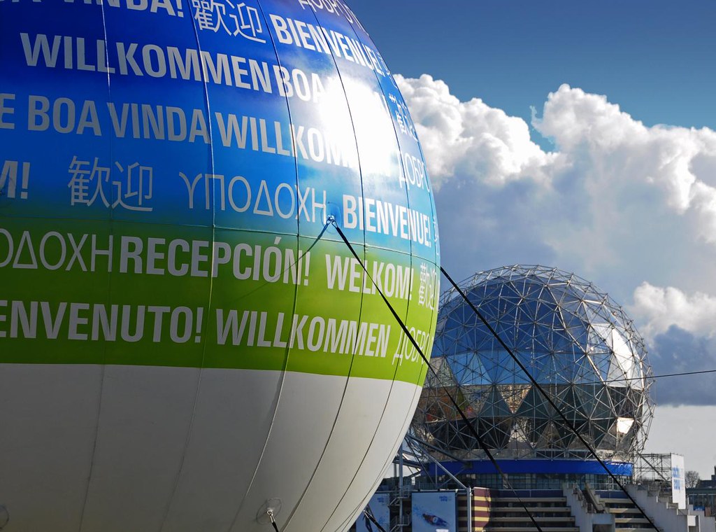 An inflatable sphere says “welcome” in many languages. Behind it is a silver spherical building.