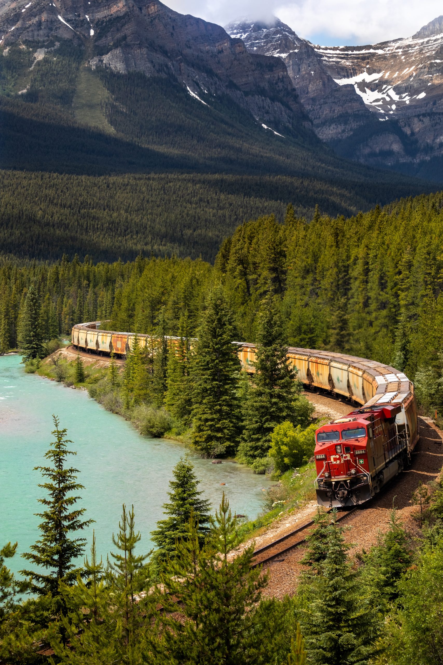 A train running through coniferous trees curves around a river beneath snowy mountains.