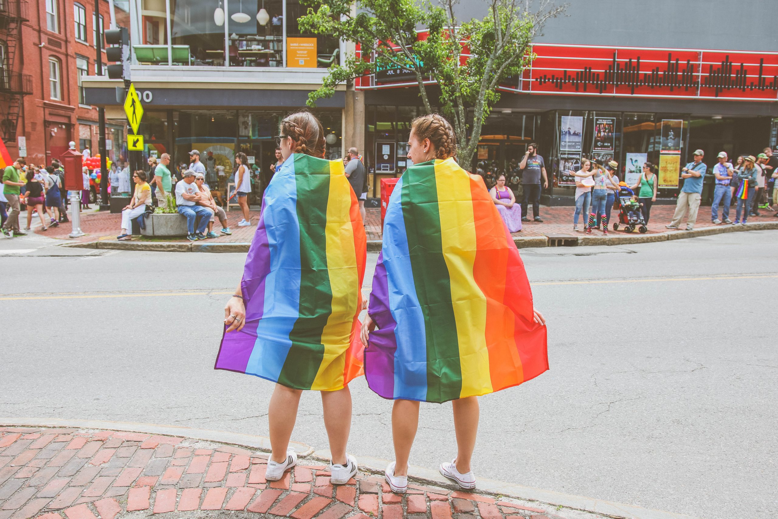 Two women wrapped in rainbow pride flags face a street with their backs to the camera.