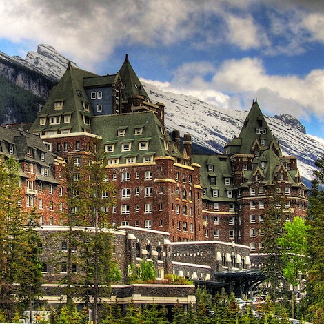 A grand hotel with brick walls and many gabled roofs in front of a snowy mountain.