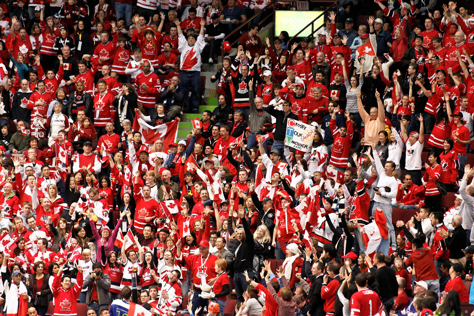 A crowd of people dressed in red and white Canadian jerseys cheer from arena seats.