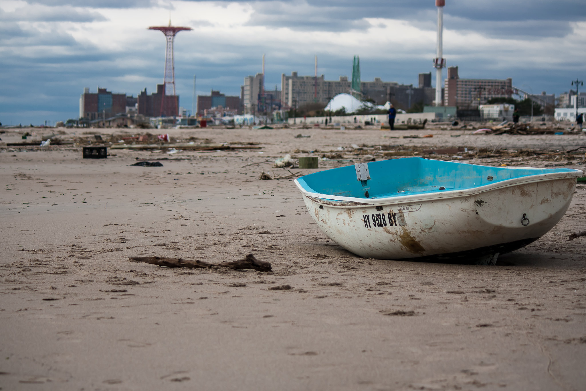 Debris and a banged-up boat are strewn across a beach. Battered buildings are in the distance.