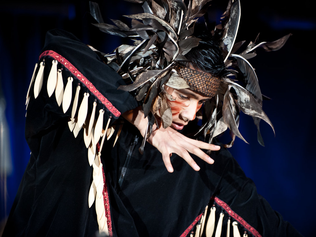 A dancer wearing a fringed black jacket, a feathered headdress, and streaks of red face paint.