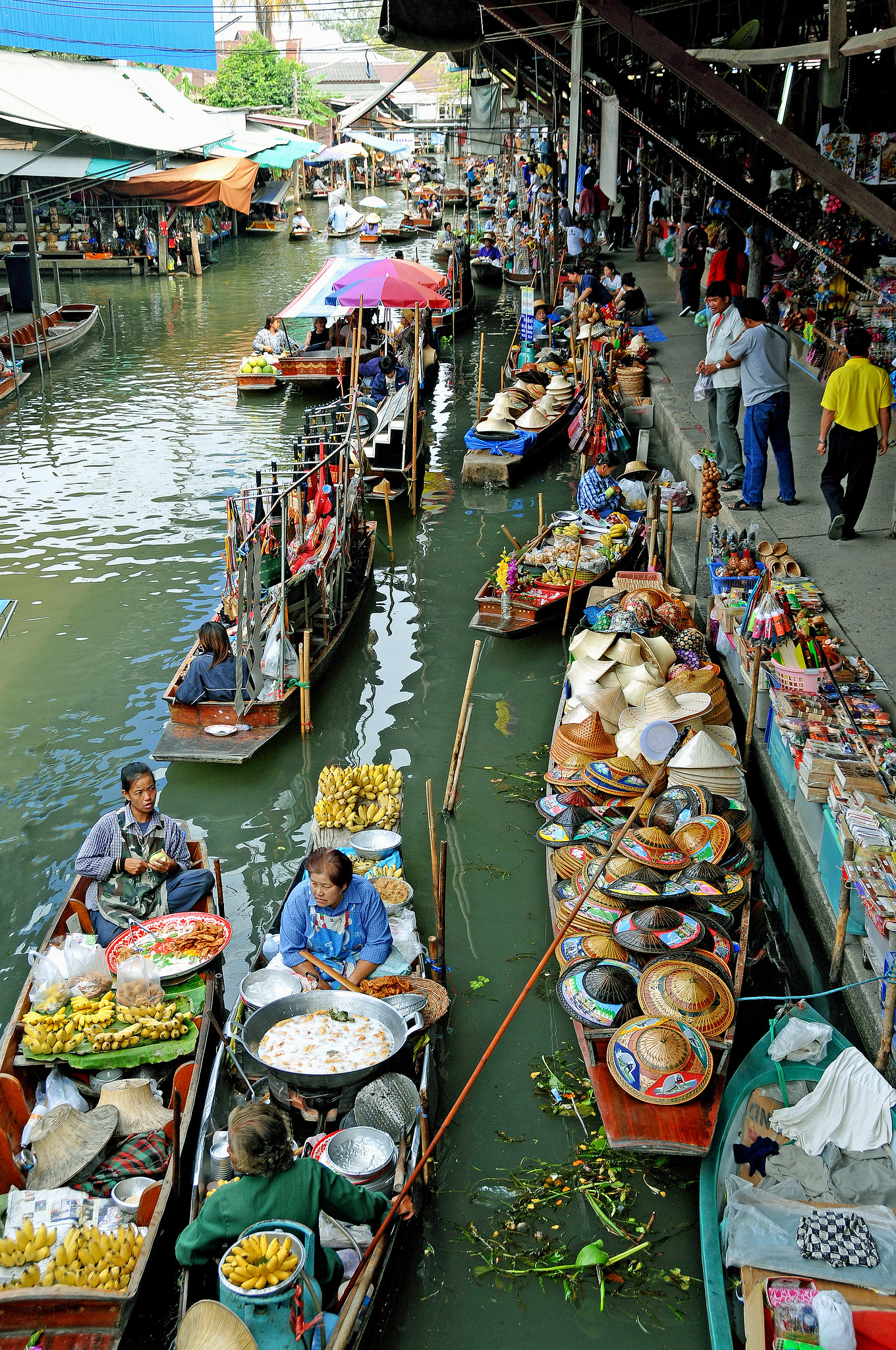 Narrow boats filled with food, clothes, hats, and much more float in a canal alongside a market.