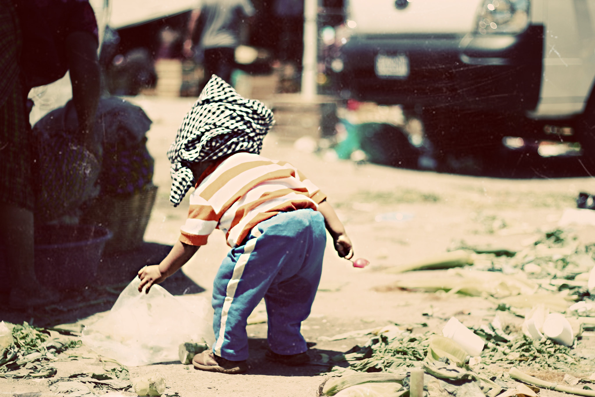 A small child with cloth wrapped around their head plays with debris in the street.