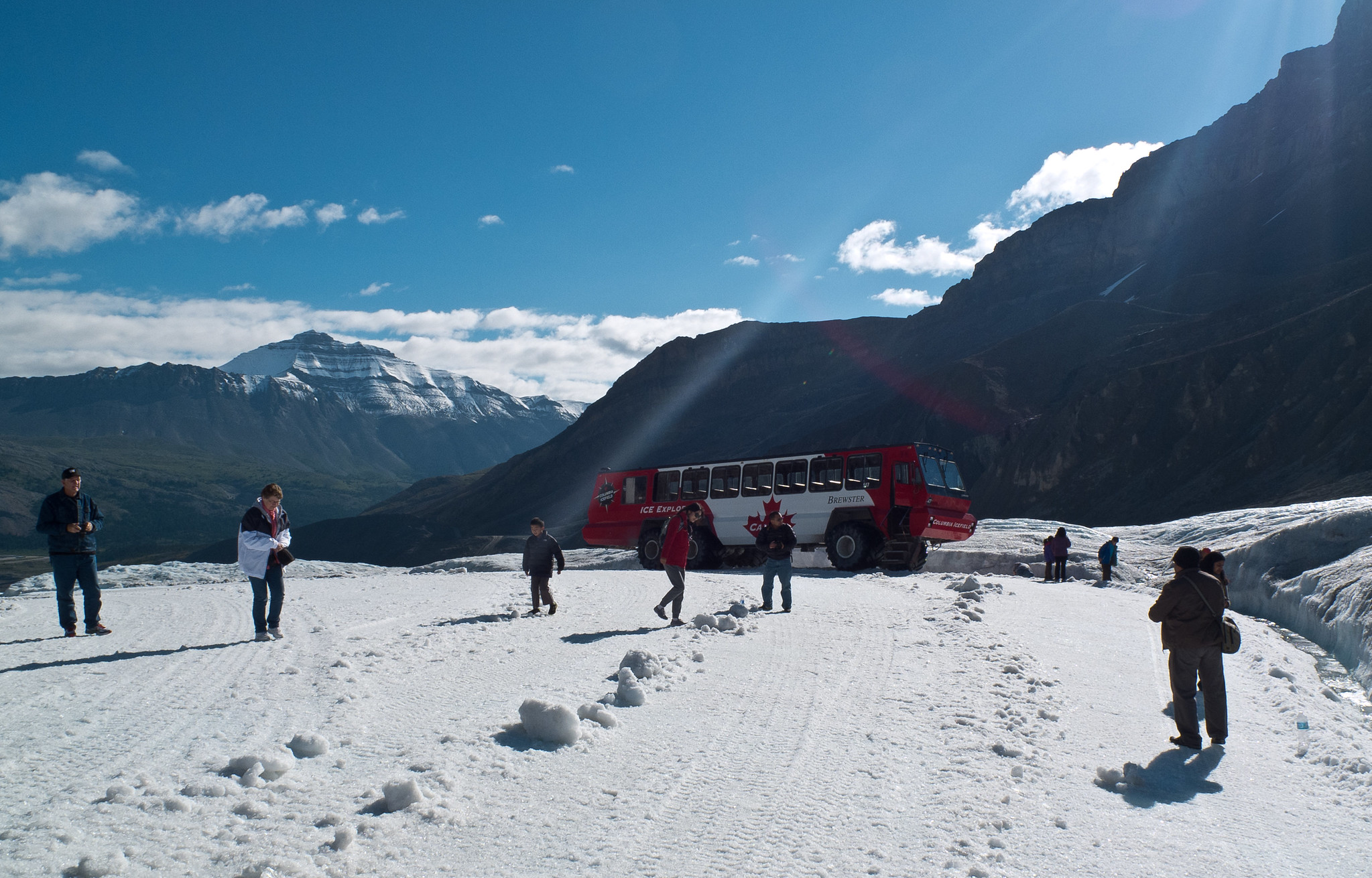 People walk across the snow in the mountains. A tour bus is parked behind them.