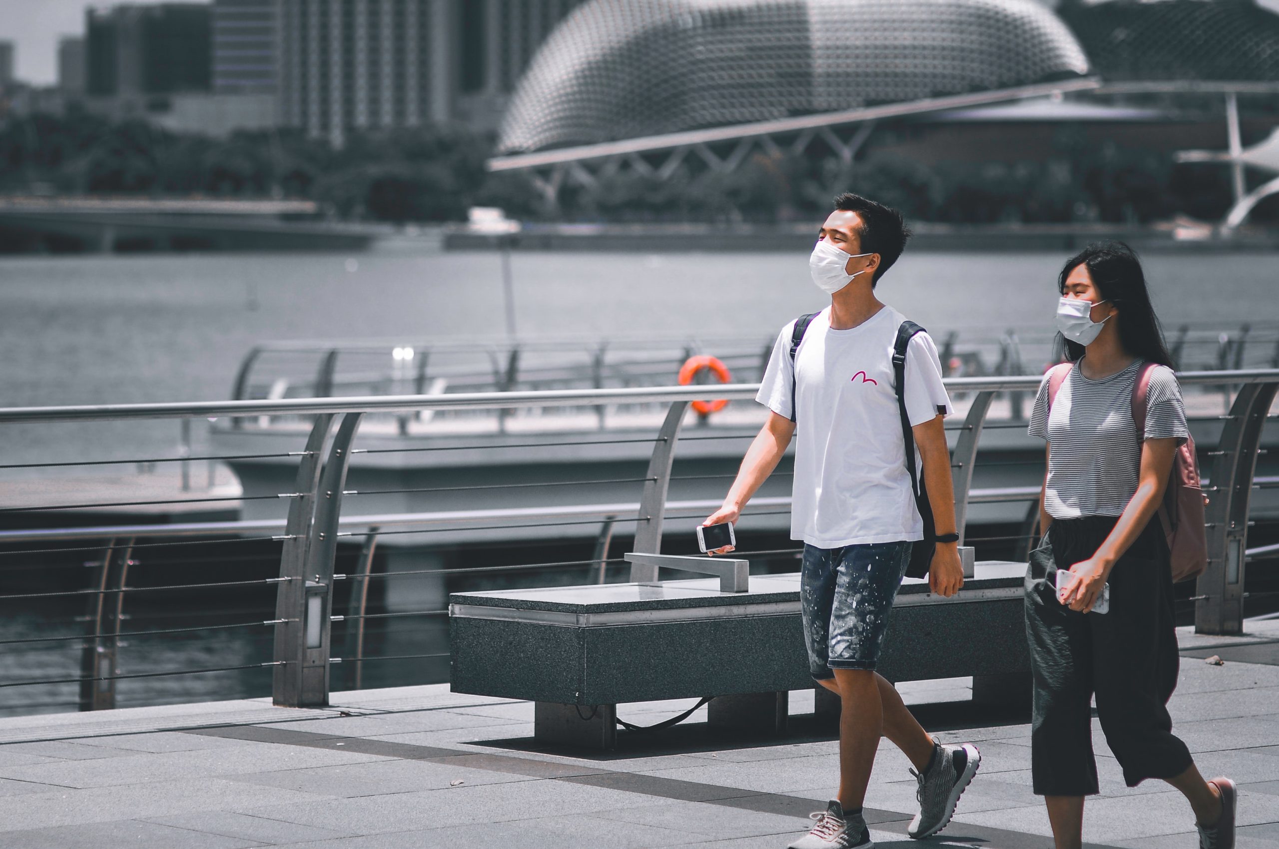 A man and woman wearing face masks and summer clothes walk past a harbour.