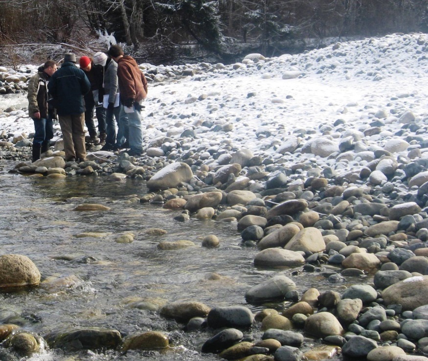 A group of peope stand on the rocky edge of a stream. There is a light dusting of snow on the ground, and large rounded rocks everywhere.