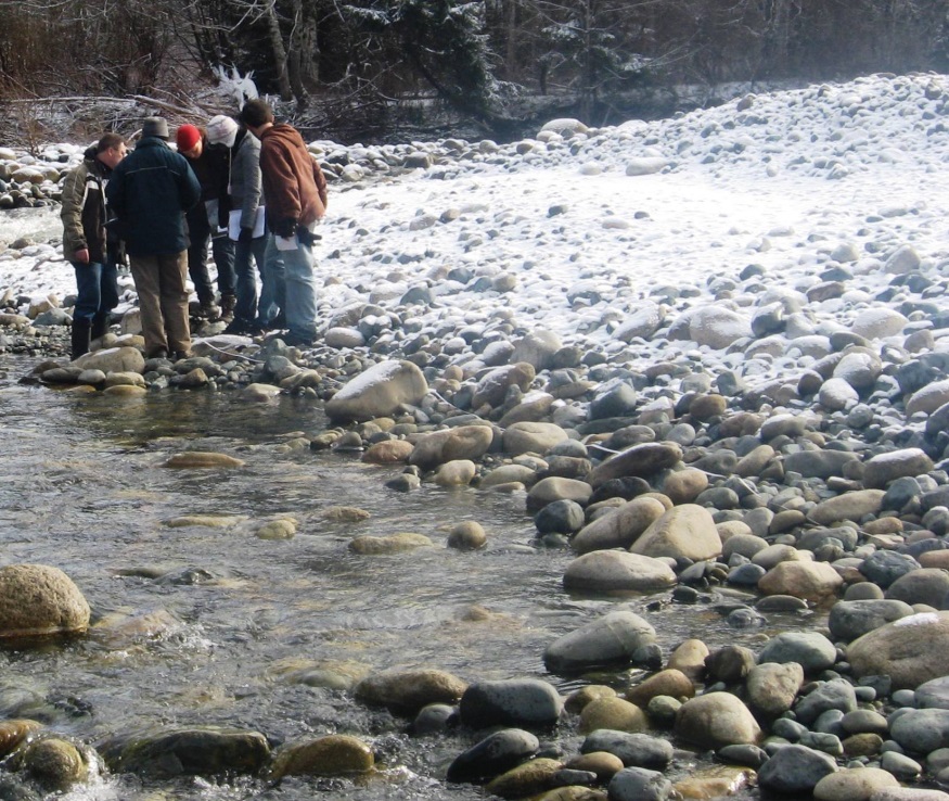 A group of people stand on the rocky edge of a stream. There is a light dusting of snow on the ground, and large rounded rocks everywhere.