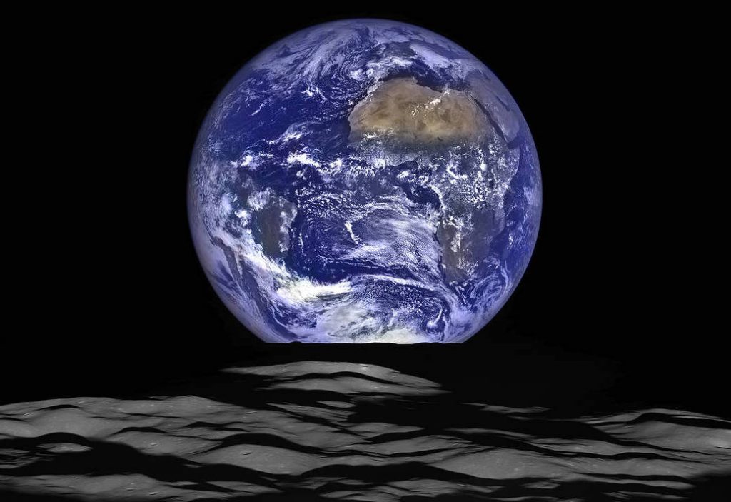 Earth appears as a deep blue sphere showing the continent of Africa. The foreground is rough terrain on the moon.
