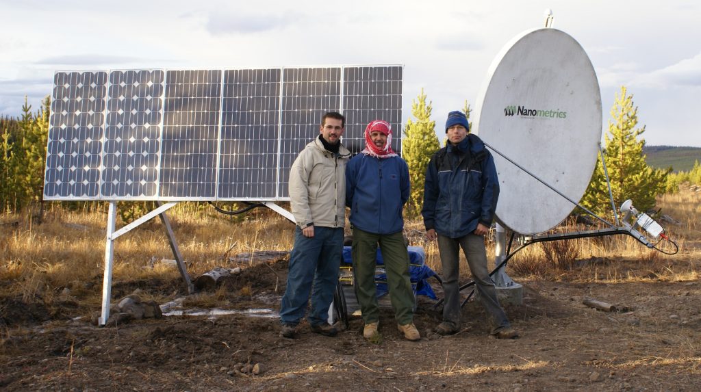 Three men stand before an array of solar panels and a satellite dish.