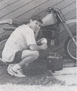 A young man crouching down in front of a motorcycle, filling it with an oil and gasoline mixture.
