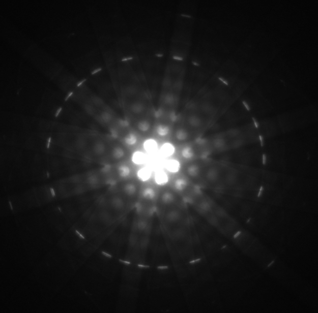 Diffraction pattern obtained for electrons diffracted by crystalline silicon is shown. The diffraction pattern has a bright spot at the center of a circle with brighter and darker regions occurring in a symmetric manner.