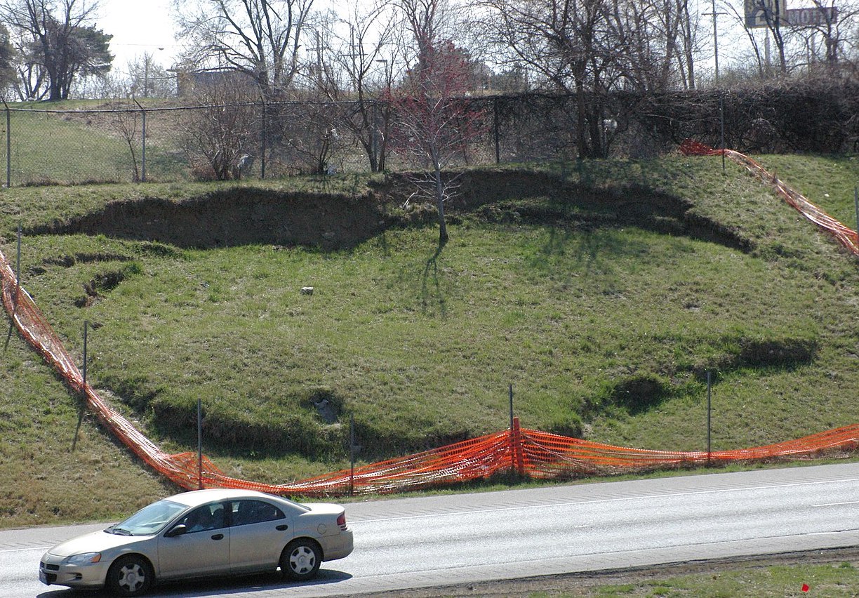 A small section of a hillside is fenced off along a road. A scarp is visible at the top of the hillside, and the hill bulges out toward the bottom.