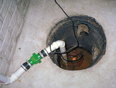 A hole in a concrete floor holds a sump pump, with piping coming out of it. There is dirty water above the pump within the hole.