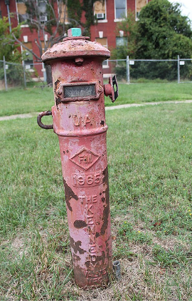 A metal post indicator valve which has a cylindrical base that sticks out of the ground and a domed top. On the side is a loop and a connector and at the top is a large screw. Information about the post indicator valve is embossed on the outside in the metal.