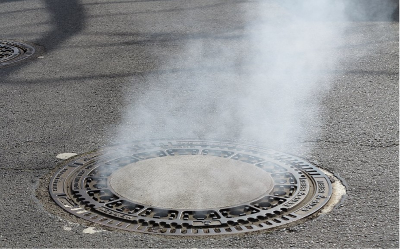 A round metal grate in the street. There is smoke rising out from underneath the grate.
