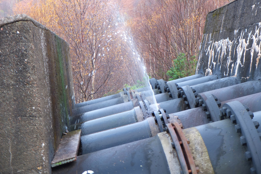 A row of six large pipes. Water is being ejected out of the side of one of the pipes.