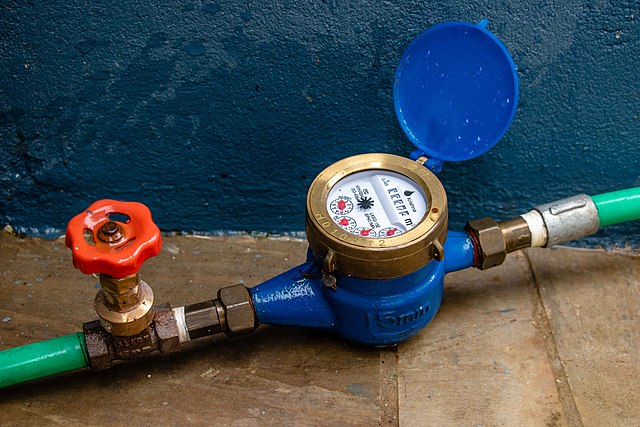 A water meter connected to a hose and a valve. The meter has a face with smaller gauges to indicate the water volume.