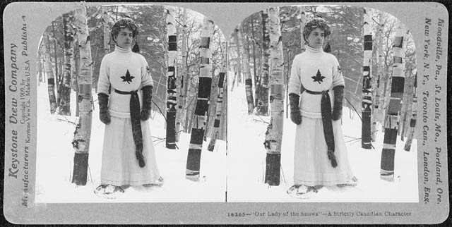 A woman in a sweater with a maple leaf, a long skirt and a toque stands in a snowy birch forest.