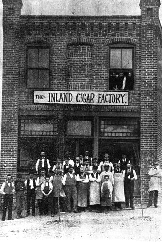 Two dozen workers in longsleeves and aprons stand outside a brick cigar factory.