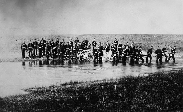 Two dozen military men cross a river, carrying weapons and pushing two cannons.
