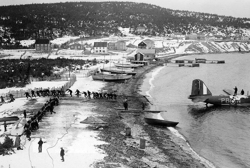Men pull an airplane out of the sea using ropes. The seaside town is covered in snow.