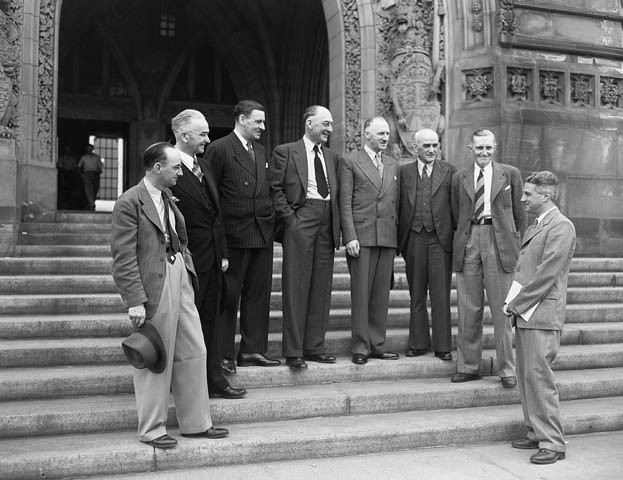 8 men in suits stand in a semicircle on the stone steps of a building, looking at the rightmost man.
