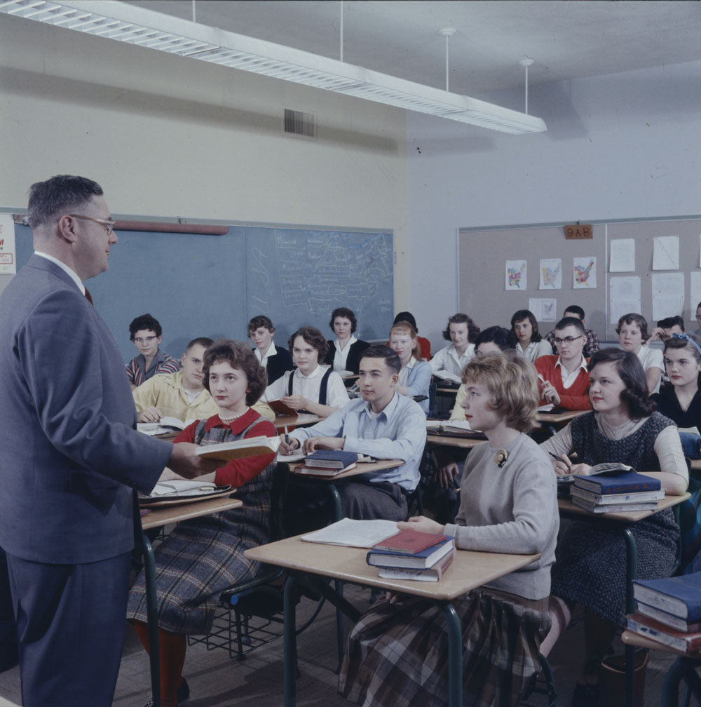A man in a suit presides over a classroom full of attentive teenagers.