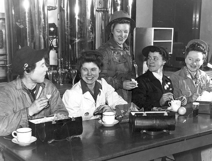 Five women in industrial uniforms sit and eat lunch out of metal lunchboxes.
