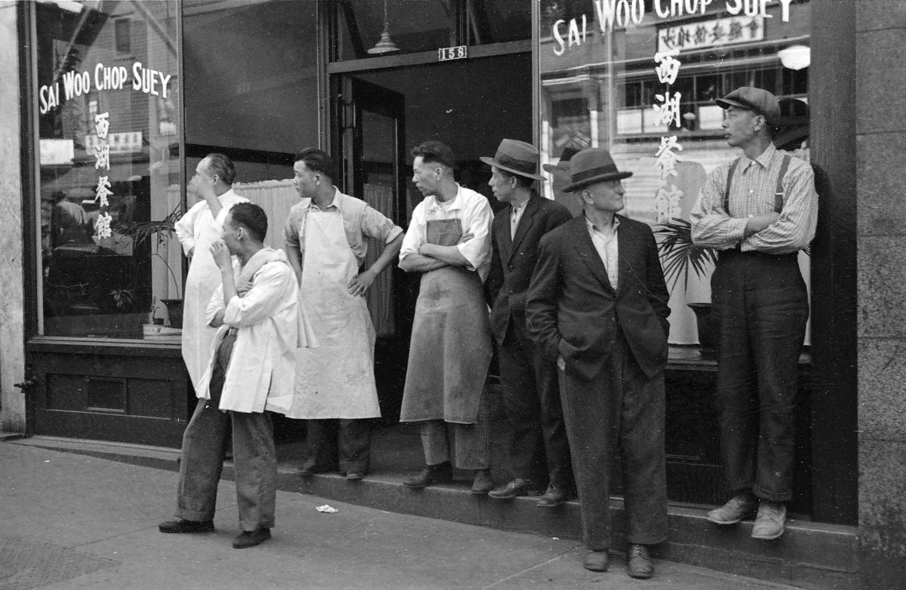 Seven men stand outside a restaurant, some dressed like kitchen workers and others in suits.