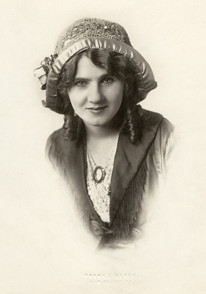 A young woman with a curl on each side of her face. She wears a large hat and has a chin dimple.