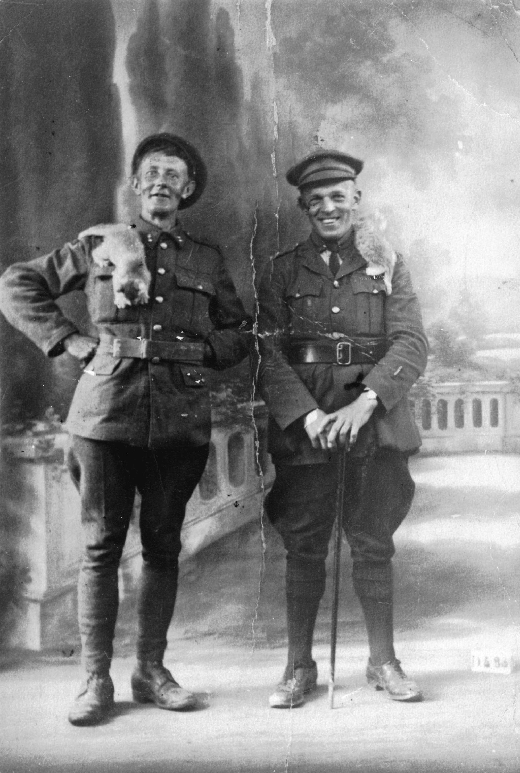 Two young men in military uniforms pose in a photo studio. Each has a cat on his shoulder.