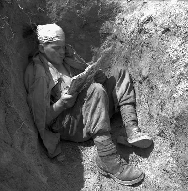 A soldier with a bandaged head sits in a foxhole, reading a comic book.