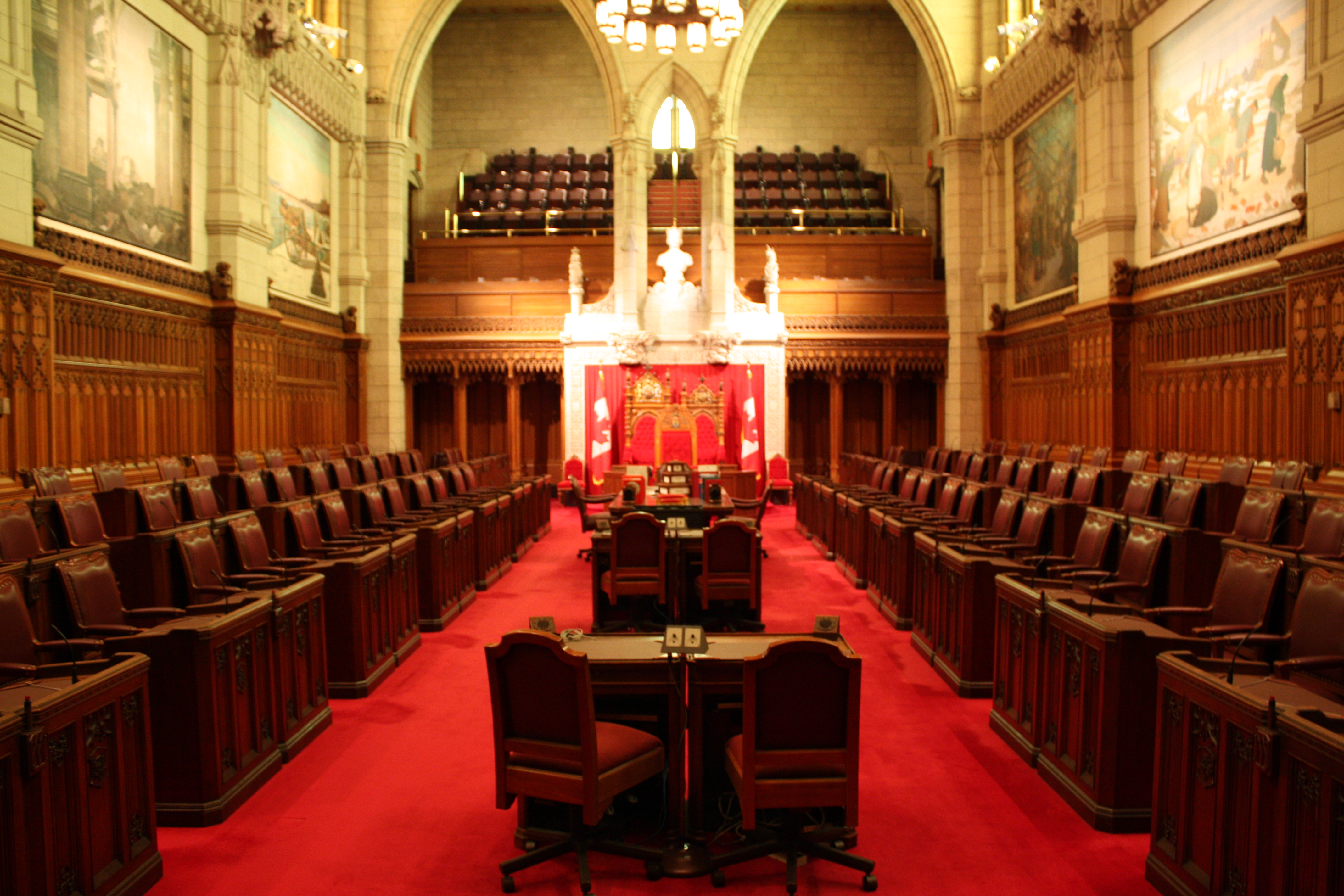 Parliamentary chamber with rows of chairs and desks, a red carpet, and a gallery.