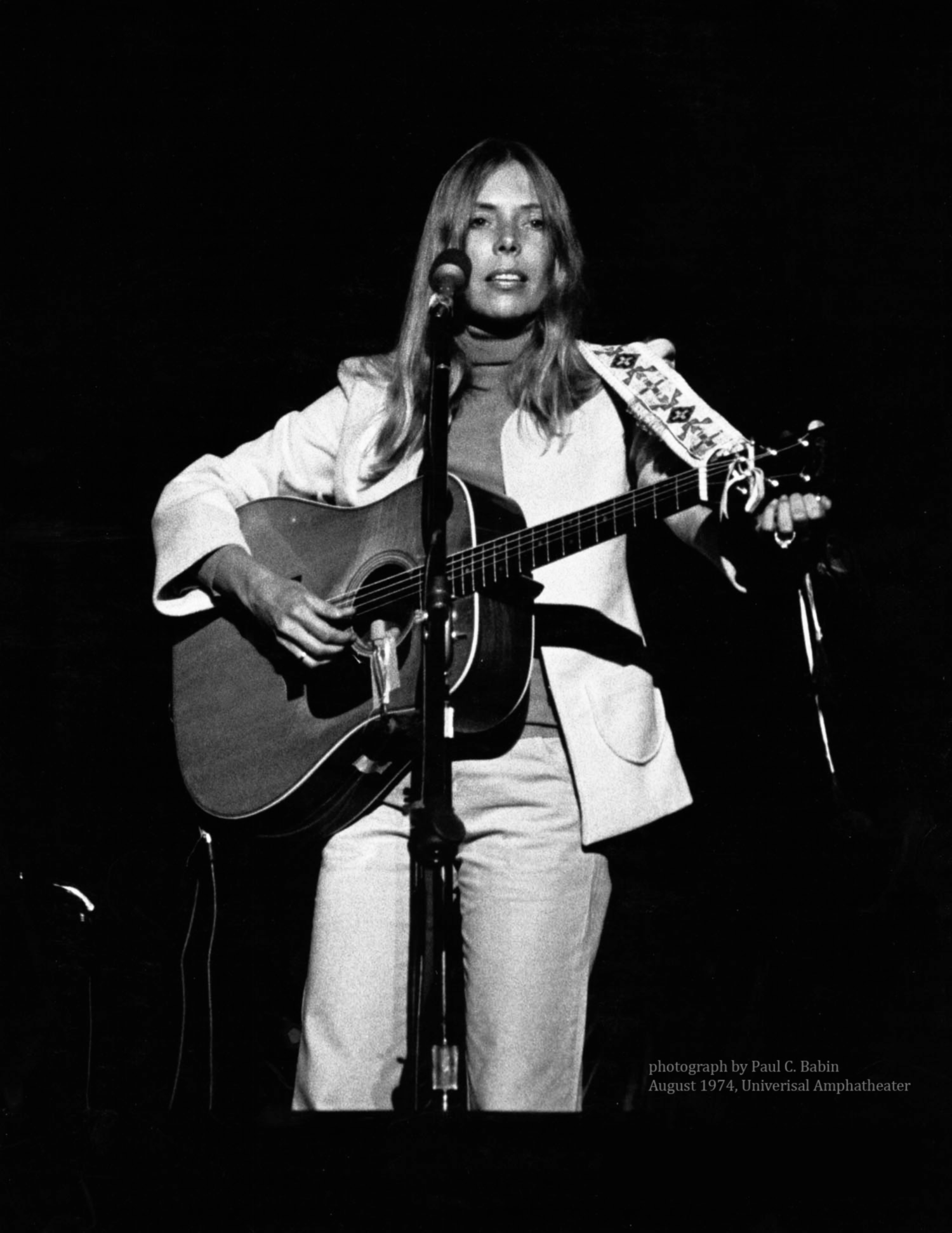 A woman stands at a microphone, holding an acoustic guitar. She wears a turtleneck and a blazer.