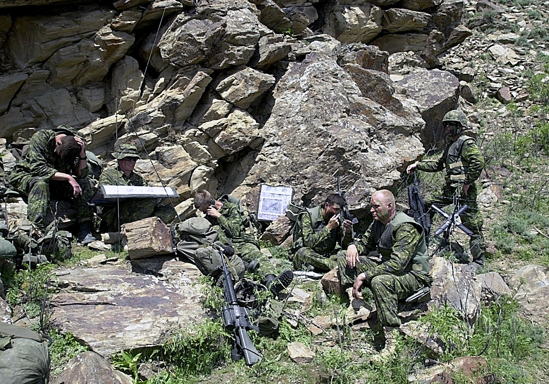 Camouflaged soldiers sit outside a cave. They hold guns and wait for something.