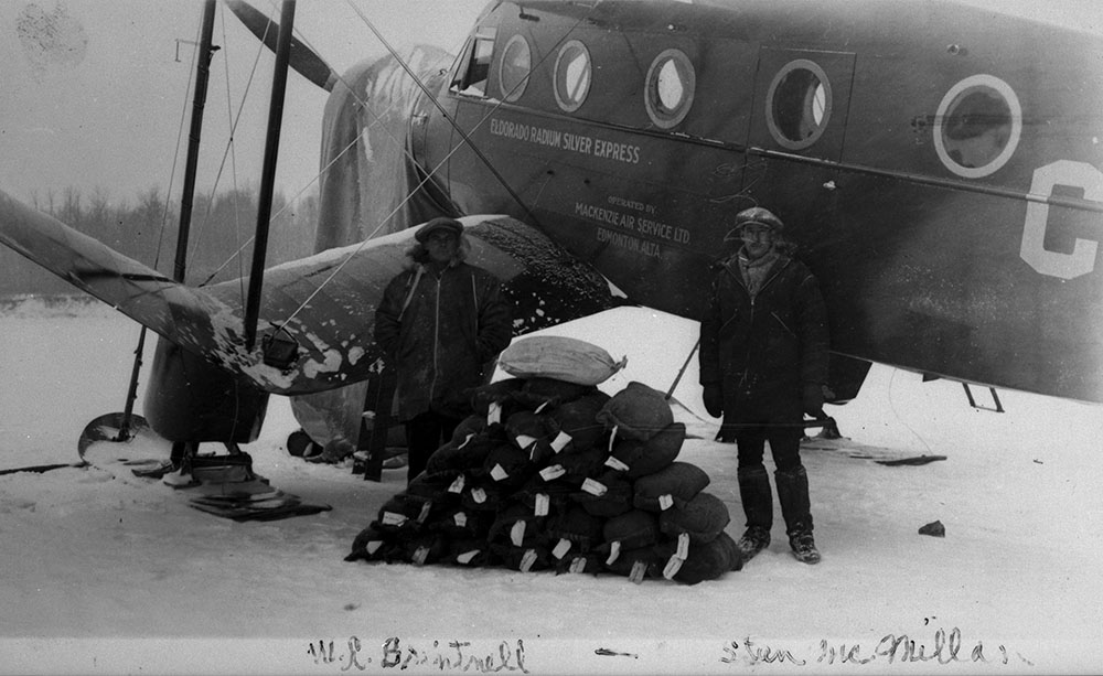 Two men stand beside a waist-high pile of small sacks. Behind them is an airplane.
