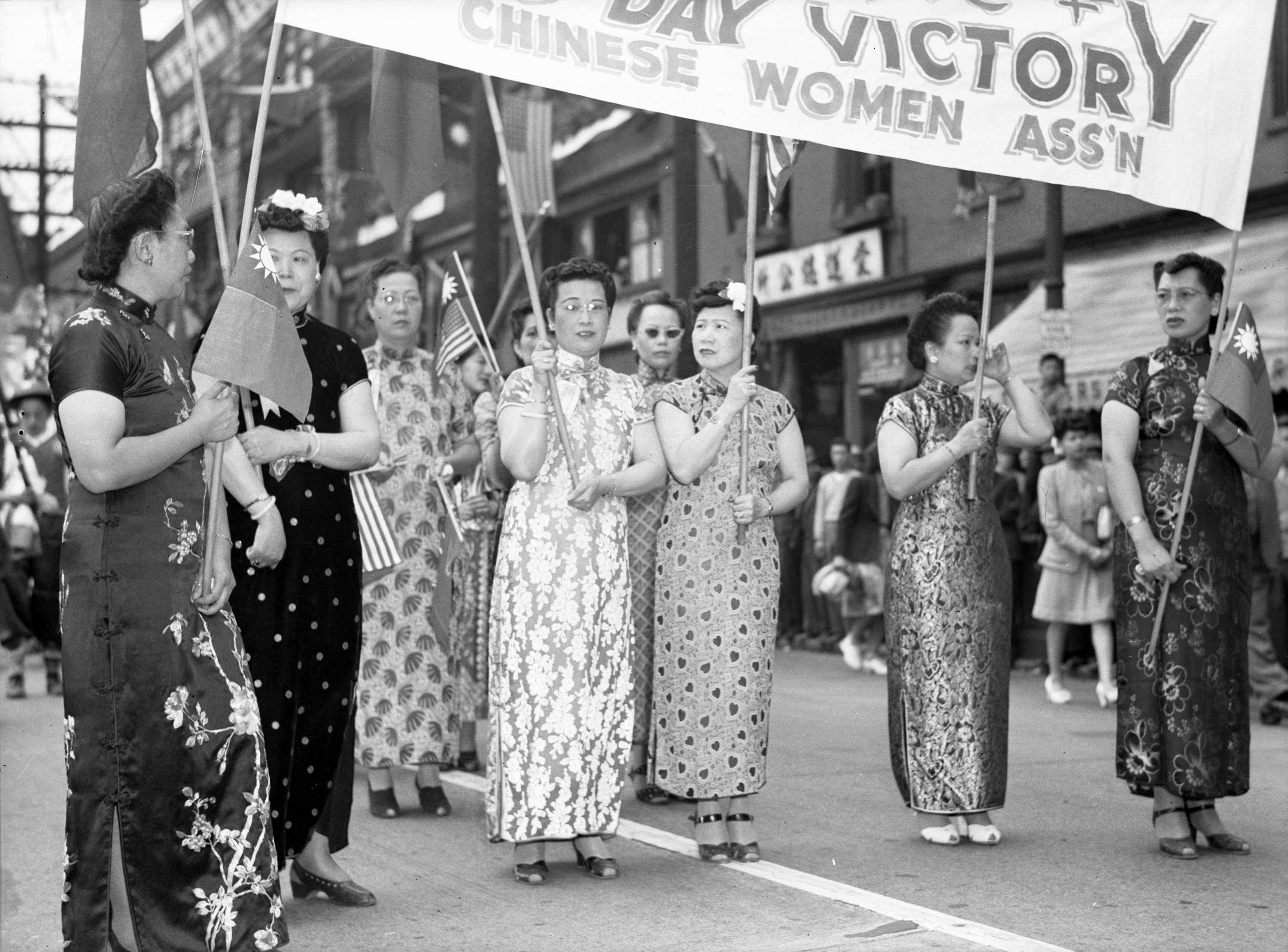 Several middle-aged women wearing qipaos walk down the street hoisting a victory banner.