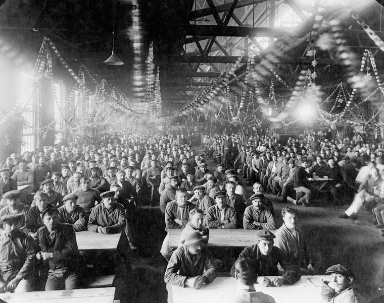 Men sit in a packed mess hall. Paper chains hang from the rafters.