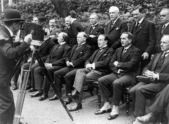 A dozen men in suits sit and stand for a photographer wearing a bowler hat.